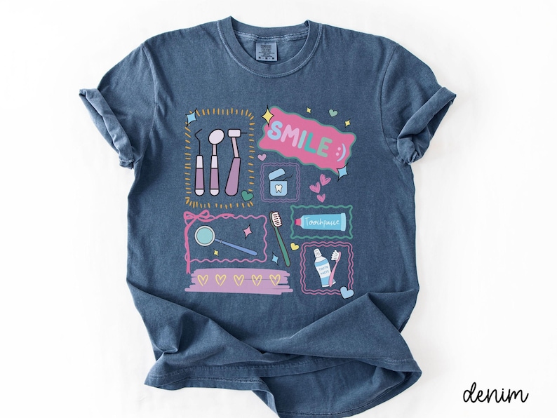 Op de afbeelding: Een denimblauw t-shirt met een grafische print van tandheelkundige instrumenten, een tandenborstel, tandpasta en flosdraad. Het ontwerp bevat de tekst "SMILE :)" in een roze tekstballon, omringd door kleine hartjes, sterretjes en decoratieve kaders.