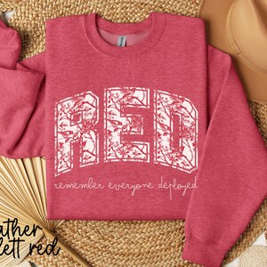 May include: Heather scarlet red sweatshirt with the word "RED" in a white floral design. The phrase "remember everyone deployed" is below the word "RED". A tan hat and woven mat are in the background.