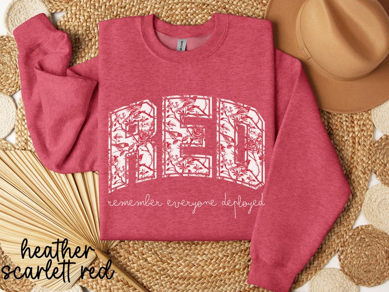May include: Heather scarlet red sweatshirt with the word "RED" in a white floral design. The phrase "remember everyone deployed" is below the word "RED". A tan hat and woven mat are in the background.