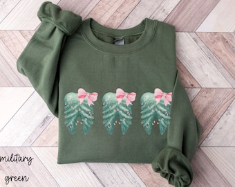 Può includere: Felpa verde militare con un design ad acquerello di tre elementi a forma di albero di Natale, ognuno ornato da un fiocco rosa. La felpa ha un girocollo e maniche lunghe con polsini a coste. Le parole "military green" sono stampate in basso.