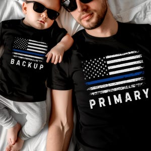 May include: A black t-shirt with a distressed American flag design and a blue line across the flag. The text "PRIMARY" is printed below the flag. A smaller black t-shirt with a similar flag design and the text "BACKUP" is printed below the flag.