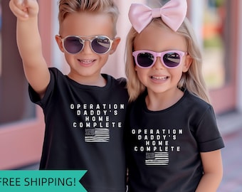 May include: Two children wearing black t-shirts with the text "Operation Daddy's Home Complete" and an American flag graphic. The girl is wearing a pink bow in her hair and both children are wearing sunglasses.