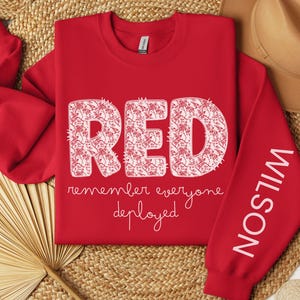 May include: A red sweatshirt with the word "RED" in large, floral-filled letters. Below, the text reads "remember everyone deployed." The right sleeve displays the name "WILSON" in white. A hat and decorative fan are in the background.