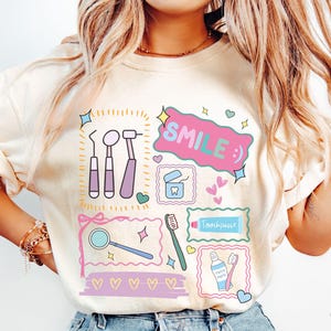 Op de afbeelding: Een ivoorkleurig t-shirt met een kleurrijke grafische print van tandheelkundige instrumenten, waaronder een spiegel, sonde, scaler, tandenborstel en tandpasta. Het ontwerp bevat de tekst "SMILE :)" in een roze tekstballon, omringd door hartjes en sterren.
