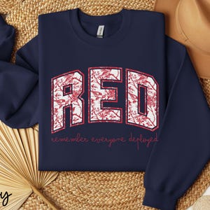 May include: Navy blue sweatshirt with the word "RED" in a white, floral-filled design. The text "remember everyone deployed" is printed below in red. A tan hat and woven mat are in the background.