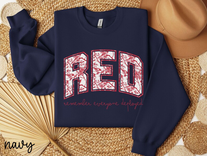 May include: Navy blue sweatshirt with the word "RED" in a white, floral-filled design. The text "remember everyone deployed" is printed below in red. A tan hat and woven mat are in the background.