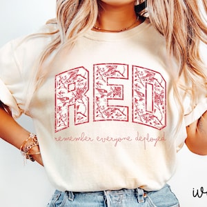 May include: Ivory-colored t-shirt with the word "RED" in a floral design. The text "remember everyone deployed" is below the word "RED". The shirt is a relaxed fit.