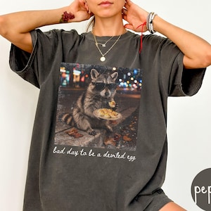 May include: A charcoal gray t-shirt featuring a graphic print of a raccoon wearing sunglasses and eating a deviled egg on a city street at night. The text below the graphic reads, bad day to be a deviled egg, in a white cursive script font.