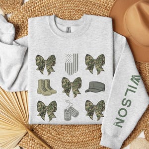 May include: Light gray sweatshirt with military-themed graphics. Features camouflage bows, boots, an American flag, a cap, and dog tags. The right sleeve displays the name "WILSON" in green lettering. The word "ash" is in the bottom left corner.