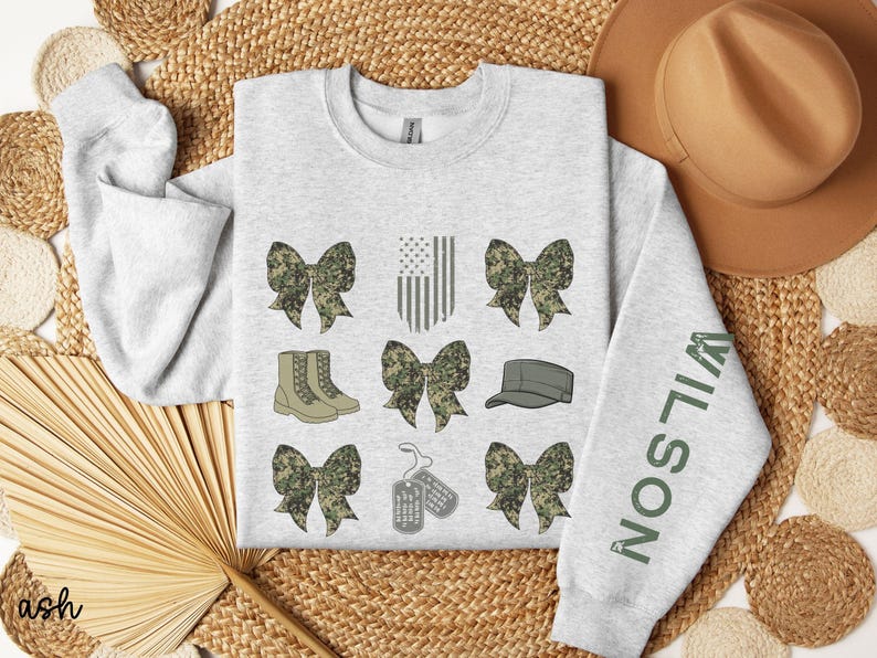 May include: Light gray sweatshirt with military-themed graphics. Features camouflage bows, boots, an American flag, a cap, and dog tags. The right sleeve displays the name "WILSON" in green lettering. The word "ash" is in the bottom left corner.