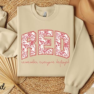 May include: Sand-colored sweatshirt with the word "RED" in large, distressed, red letters filled with a floral pattern. Below the word "RED" is the phrase "remember everyone deployed" in a smaller, red font. The sweatshirt has long sleeves.
