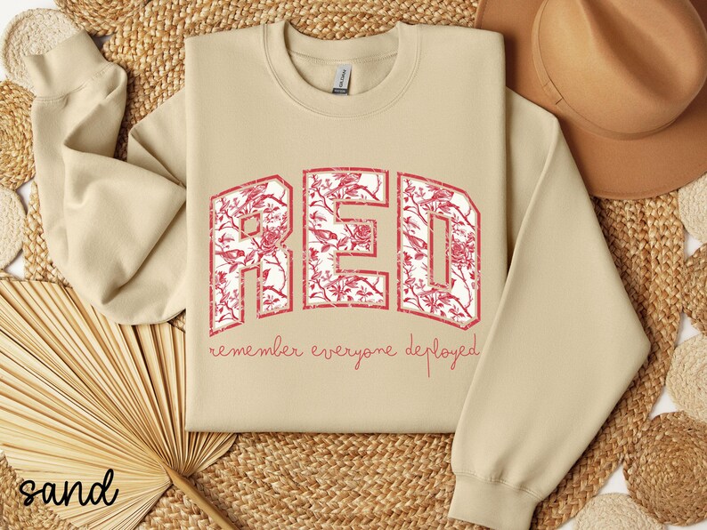 May include: Sand-colored sweatshirt with the word "RED" in large, distressed, red letters filled with a floral pattern. Below the word "RED" is the phrase "remember everyone deployed" in a smaller, red font. The sweatshirt has long sleeves.