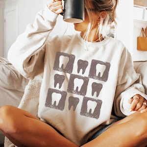 K&ouml;nnte beinhalten: Ein cremefarbenes Sweatshirt mit einem Muster aus Z&auml;hnen in grauen Quadraten. Das Sweatshirt hat eine lockere Passform und einen Rundhalsausschnitt. Das Bild zeigt eine Person, die eine schwarze Tasse h&auml;lt.