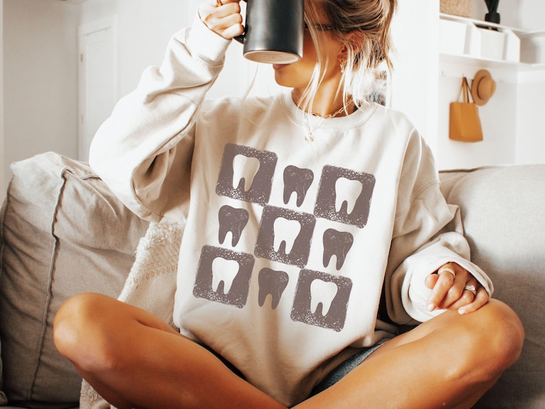 K&ouml;nnte beinhalten: Ein cremefarbenes Sweatshirt mit einem Muster aus Z&auml;hnen in grauen Quadraten. Das Sweatshirt hat eine lockere Passform und einen Rundhalsausschnitt. Das Bild zeigt eine Person, die eine schwarze Tasse h&auml;lt.