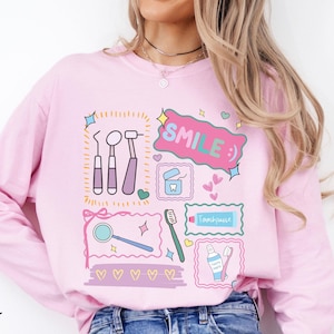 Op de afbeelding: Een lichtroze sweatshirt met lange mouwen en een grafische print van tandheelkundige instrumenten, waaronder een spiegel, tandenborstel, tandpasta en flosdraad. Het ontwerp bevat de tekst "SMILE :)" in een roze tekstballon, omringd door hartjes.
