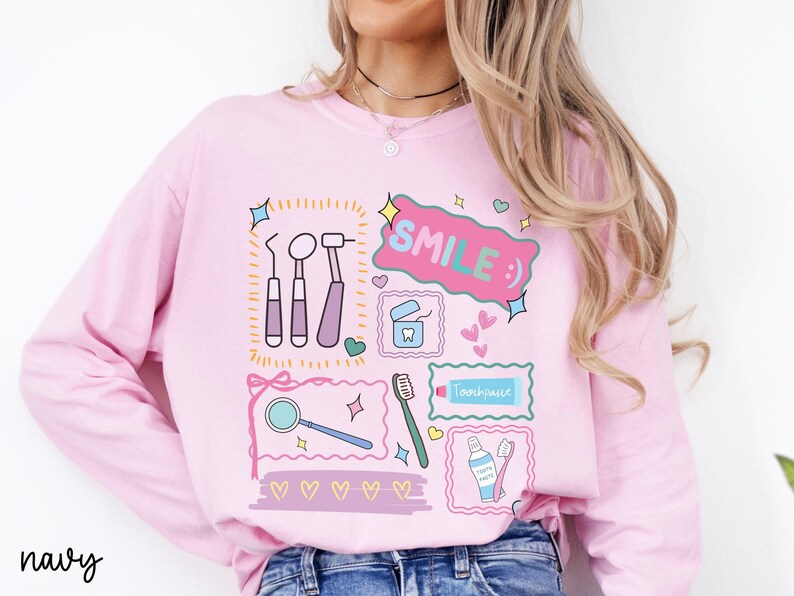 Op de afbeelding: Een lichtroze sweatshirt met lange mouwen en een grafische print van tandheelkundige instrumenten, waaronder een spiegel, tandenborstel, tandpasta en flosdraad. Het ontwerp bevat de tekst "SMILE :)" in een roze tekstballon, omringd door hartjes.