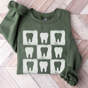 K&ouml;nnte beinhalten: Gr&uuml;nes Sweatshirt mit einem wei&szlig;en Zahnmotiv, das in einem Rastermuster wiederholt wird. Die Z&auml;hne befinden sich in einem quadratischen Rahmen mit einem Used-Look. Das Sweatshirt hat einen Rundhalsausschnitt und lange &Auml;rmel.