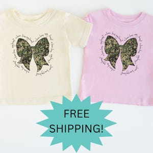 Peut inclure: Deux t-shirts pour tout-petits avec un n&oelig;ud camouflage et le texte "Mon c&oelig;ur a un h&eacute;ros, mon h&eacute;ros a mon c&oelig;ur".