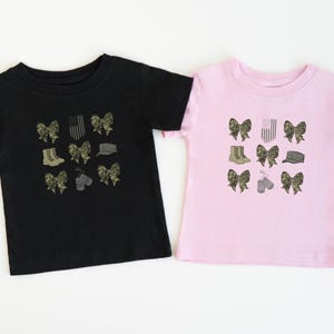 May include: Two t-shirts, one black and one pink, with a design of camouflage bows, boots, a hat, dog tags, and an American flag.