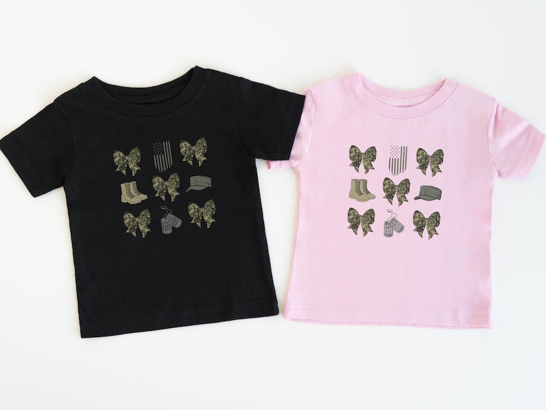 May include: Two t-shirts, one black and one pink, with a design of camouflage bows, boots, a hat, dog tags, and an American flag.