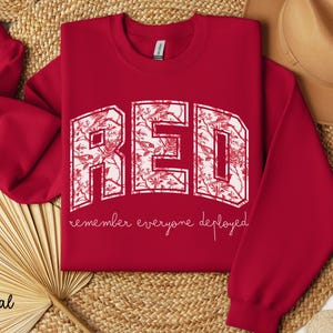 May include: A cardinal red sweatshirt with the word "RED" in large, white, floral-patterned letters. Below the letters, the phrase "remember everyone deployed" is written in a cursive font. The sweatshirt is a crew neck style.