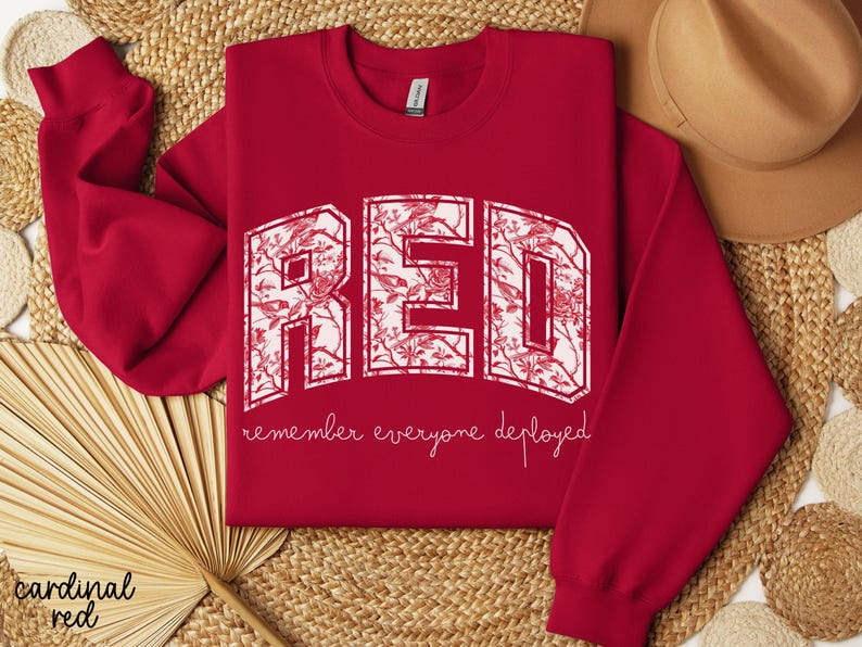 May include: A cardinal red sweatshirt with the word "RED" in large, white, floral-patterned letters. Below the letters, the phrase "remember everyone deployed" is written in a cursive font. The sweatshirt is a crew neck style.