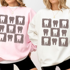 K&ouml;nnte beinhalten: Zwei Sweatshirts, eines rosa und eines wei&szlig;, jeweils mit einem sich wiederholenden Muster aus Z&auml;hnen in quadratischen Rahmen. Die Z&auml;hne sind in einem ged&auml;mpften Braun gehalten, und die Sweatshirts haben einen Rundhalsausschnitt.
