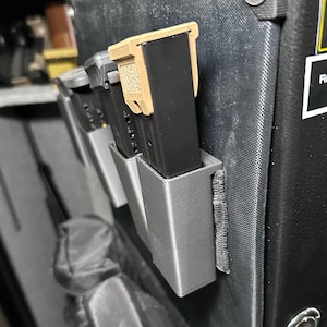May include: A black gun safe with a gray plastic magazine holder attached to the inside. The magazine holder has two black magazines inside. The safe has a black handle and a black button.