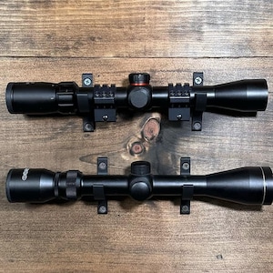 May include: Two black rifle scopes mounted on a wooden surface. The top scope has a red adjustment knob and a set of rings. The bottom scope has a silver band around the lens.