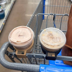 May include: Two disposable coffee cups with lids in a shopping cart. The cup on the left has a brown lid and the cup on the right has a clear lid. Both cups have brown paper sleeves with a logo that says "SCOUTS".