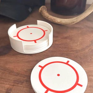 May include: White coasters with a red target design. The coasters are stacked in a white holder, with one coaster separate. A black cap and a brown glass are in the background.