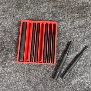 May include: A set of eight black metal punch tools in a red plastic case. The tools are arranged in order of size, with the largest tool on the left and the smallest tool on the right. The tools are labeled with their sizes in inches.