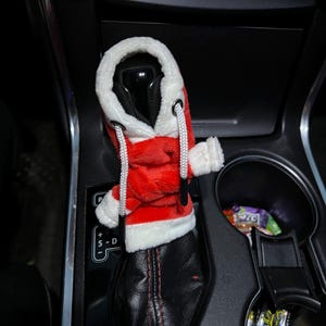 Puede incluir: Una funda de palanca de cambios de felpa roja y blanca con tem&aacute;tica de Pap&aacute; Noel para un autom&oacute;vil. La funda tiene forma de sudadera con capucha con ribete blanco y un cord&oacute;n blanco.