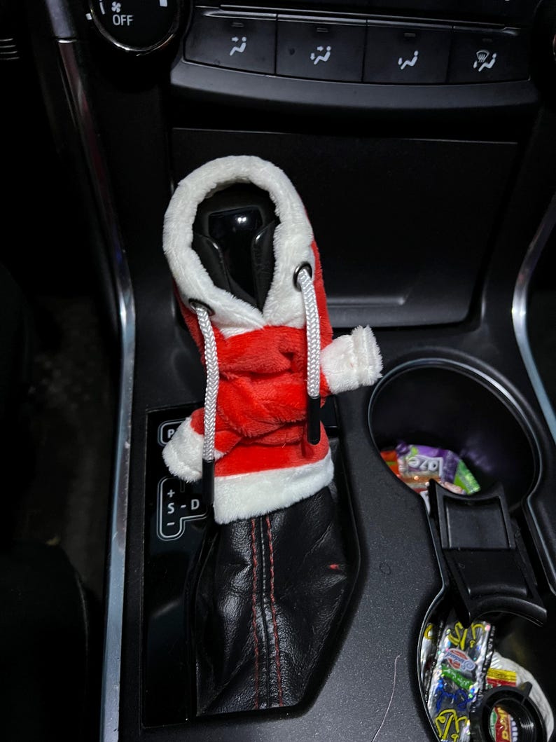 Puede incluir: Una funda de palanca de cambios de felpa roja y blanca con tem&aacute;tica de Pap&aacute; Noel para un autom&oacute;vil. La funda tiene forma de sudadera con capucha con ribete blanco y un cord&oacute;n blanco.
