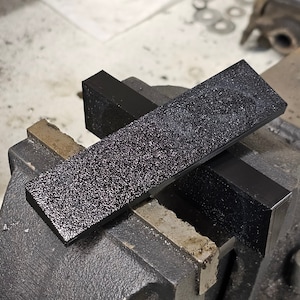 May include: A black textured rectangular block clamped in a metal vise. The block has a rough, grainy surface.