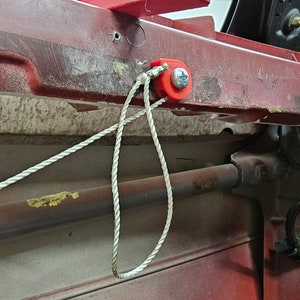 May include: A close-up of a red metal frame with a white rope attached to a red plastic clip. The rope is threaded through a hole in the frame and secured with a screw.