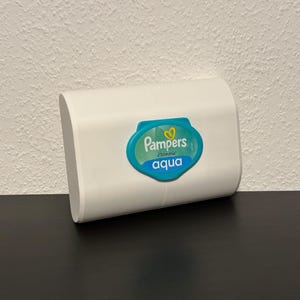 Op de afbeelding: Witte plastic container met babydoekjes met een blauw en groen label waarop "Pampers" en "Aqua" in witte letters staat.