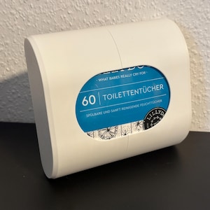 Op de afbeelding: Een witte, gebogen, rechthoekige container met een blauw ovaal venster met de tekst "60 TOILETTENTÜCHER". De container heeft een gladde afwerking en staat op een zwart oppervlak.