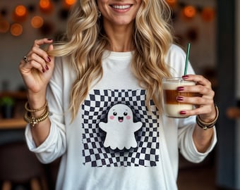 Pode incluir: Camiseta branca de manga comprida com um design xadrez em preto e branco com um fantasma de desenho animado com um rosto sorridente. O fantasma está no centro do design xadrez. A camiseta está sendo usada por uma pessoa que está segurando um copo de café gelado com um canudo verde.