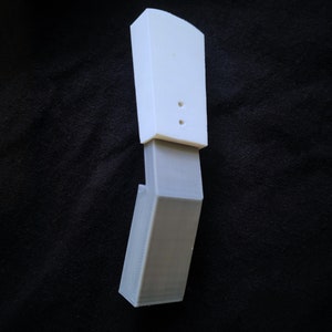 Puede incluir: Un soporte para teléfono impreso en 3D blanco y gris. El soporte tiene una base rectangular y una parte superior curva. La parte superior tiene dos pequeños agujeros.