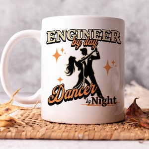 May include: White ceramic mug with a black and orange design. The mug features the text "Engineer by day Dancer by Night" with a silhouette of a couple dancing.