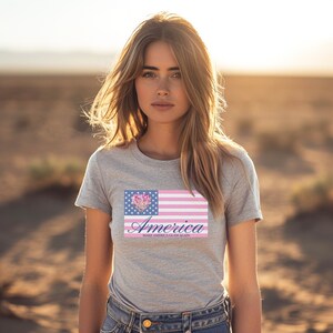 May include: A gray t-shirt featuring a stylized American flag design. The flag incorporates a floral heart and pink stripes, with the word "America" in cursive and the phrase "Make America Glam Again" below.