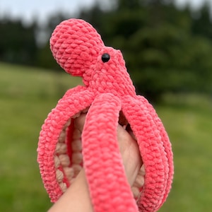 May include: A pink crocheted octopus plush toy with black eyes. The octopus is being held by a hand.
