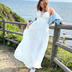 May include: A woman wearing a white maxi dress with a denim jacket draped over her shoulders. She is standing on a wooden walkway with a view of the ocean in the background.