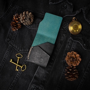 Puede incluir: Una cartera de cuero negro con interior turquesa, colocada sobre una chaqueta vaquera negra. La cartera tiene costuras blancas. Llaves doradas, un reloj de bolsillo y pi&ntilde;as se disponen como elementos decorativos.