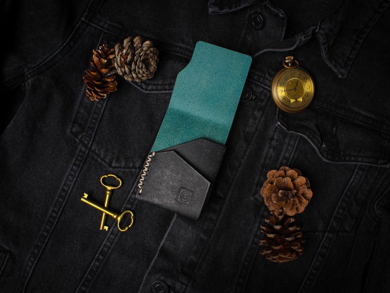 Puede incluir: Una cartera de cuero negro con interior turquesa, colocada sobre una chaqueta vaquera negra. La cartera tiene costuras blancas. Llaves doradas, un reloj de bolsillo y pi&ntilde;as se disponen como elementos decorativos.