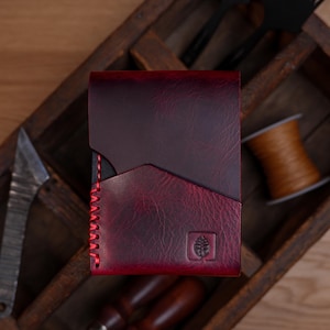May include: A deep burgundy leather wallet featuring a textured finish, a diagonal pocket design, and bright red stitching along the side. A small square logo depicting a stylized leaf is embossed on the bottom right corner of the wallet.