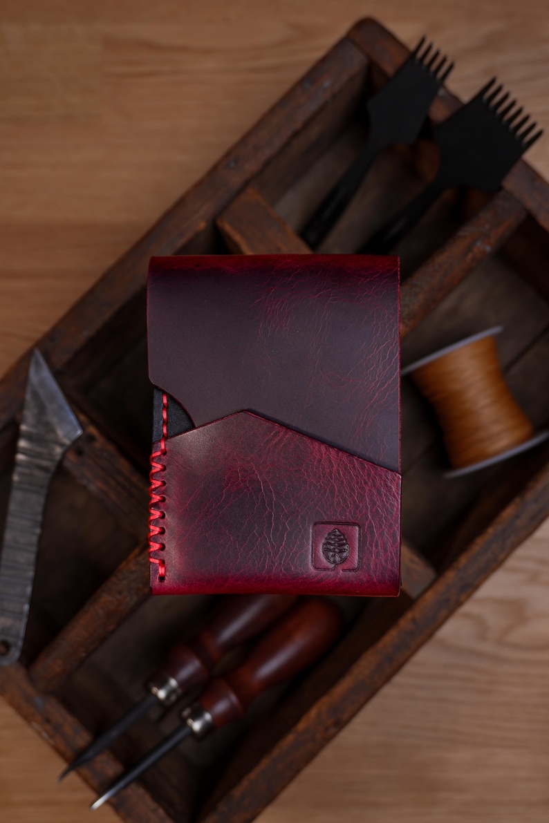 May include: A deep burgundy leather wallet featuring a textured finish, a diagonal pocket design, and bright red stitching along the side. A small square logo depicting a stylized leaf is embossed on the bottom right corner of the wallet.