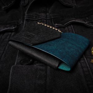 Puede incluir: Una cartera plegable con interior turquesa y exterior negro, apoyada sobre una chaqueta vaquera negra. La cartera tiene una solapa gris oscuro con detalles cosidos. Dos llaves antiguas de lat&oacute;n est&aacute;n junto a la cartera.