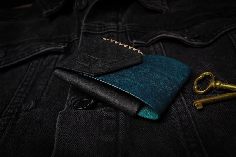 Puede incluir: Una cartera plegable con interior turquesa y exterior negro, apoyada sobre una chaqueta vaquera negra. La cartera tiene una solapa gris oscuro con detalles cosidos. Dos llaves antiguas de lat&oacute;n est&aacute;n junto a la cartera.